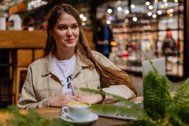 Kafkasyalı romantik kadın blogcu kahve içerek rahatlıyor, klasik kafede oturuyor, kadın serbest çalışıyor yeni fikirleri düşünüyor, dizüstü bilgisayarda çalışıyor, tuğla duvar, eski mobilyalar, başka tarafa bakıyor.