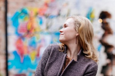 Güzel, düşünceli kadın sırıtışları, parlayan gözleri, beyaz tenli genç bir kadın, gözlerini kaçıran sarışın kız, gri ceket ve bej elbise, sokak fotoğrafı, klasik geçmiş, düşünceli bir ifade.