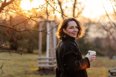 Güzel, genç, beyaz bir kadın güneşli bir bahar gününün, parkın, kiraz bahçesinin, romantik bir portrenin, kahvenin, siyah ceketin ve atkının, kıvırcık kahverengi saçların, zarif bardakların, gün batımının ve güneşin tadını çıkar.