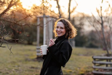 Güzel, genç, beyaz bir kadın güneşli bir bahar gününün, parkın, kiraz bahçesinin, romantik bir portrenin, kahvenin, siyah ceketin ve atkının, kıvırcık kahverengi saçların, zarif bardakların, gün batımının ve güneşin tadını çıkar.