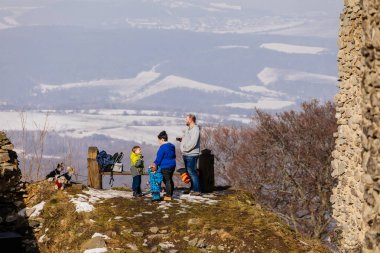 Aile bankta, anne baba çay, panoramik manzara, ortaçağ gotik kalesi Kostomlaty veya Sukoslav, kar, tepede taş yıkıntısı, güneşli kış günü, Çek Cumhuriyeti, 20 Şubat 2021
