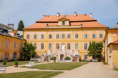 Krasny Dvur Chateau, Kuzey Bohemya, Çek Cumhuriyeti, 19 Haziran 2021: Merdivenleri olan barok sarı kale, yeşil çimenli ön bahçe ve güneşli yaz gününde beyaz banklar.