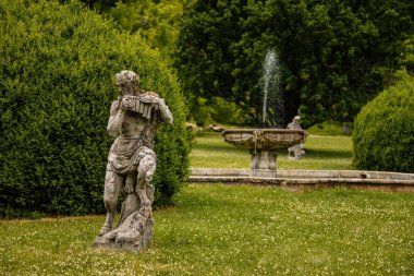 Valec, Batı Bohemya, Çek Cumhuriyeti, 19 Haziran 2021: Uzun ağaçlı ve yeşil çimenli İngiliz parkı, taş kase etrafındaki tanrı heykelleri ve satirlerle güneşli yaz günü şatonun yanındaki barok çeşmesi..