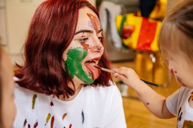 Aile eğlencesi ev konsepti. Anne ve küçük kızı resim çiziyor, boya fırçaları ve suluboya boya kullanarak eğleniyorlar. Çocuklar ve ebeveynler birlikte resim yapıyor, yüzlerinde suluboya, ev karantinası.