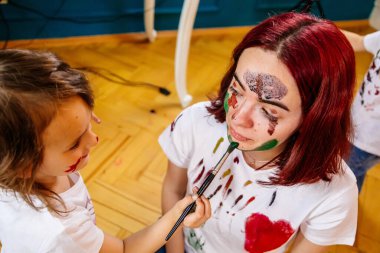 Aile eğlencesi ev konsepti. Anne ve küçük kızı resim çiziyor, boya fırçaları ve suluboya boya kullanarak eğleniyorlar. Çocuklar ve ebeveynler birlikte resim yapıyor, yüzlerinde suluboya, ev karantinası.