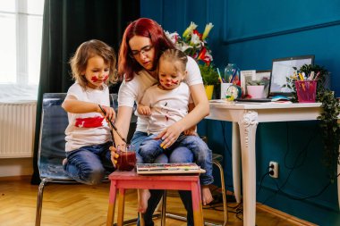 Aile hobileri boş zaman aktiviteleri ev konsepti. Anne ve küçük kızı, boya fırçaları ve suluboya boyalarla eğlenerek resim çiziyorlar. Çocuklar ve ebeveynler birlikte resim yapıyorlar, vücut boyası, ev karantinası..