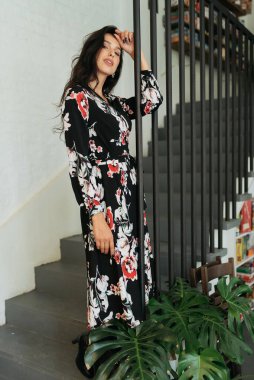 Genç zarif kız içeride poz veriyor. Duvarda ve pencerede zarif elbiseler içinde güzel bir iş kadını.