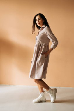 Genç zarif kız içeride poz veriyor. Duvarda ve pencerede zarif elbiseler içinde güzel bir iş kadını.