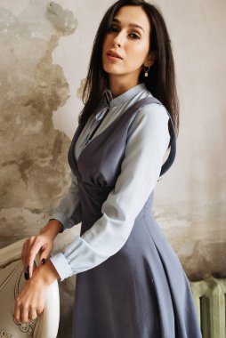 Genç zarif kız içeride poz veriyor. Duvarda ve pencerede zarif elbiseler içinde güzel bir iş kadını.