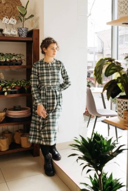 Genç zarif kız içeride poz veriyor. Duvarda ve pencerede zarif elbiseler içinde güzel bir iş kadını.