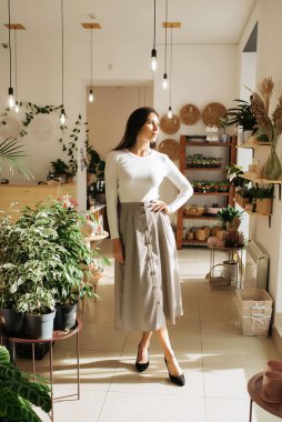 Genç zarif kız içeride poz veriyor. Duvarda ve pencerede zarif elbiseler içinde güzel bir iş kadını.