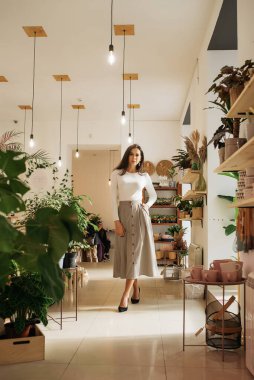 Genç zarif kız içeride poz veriyor. Duvarda ve pencerede zarif elbiseler içinde güzel bir iş kadını.