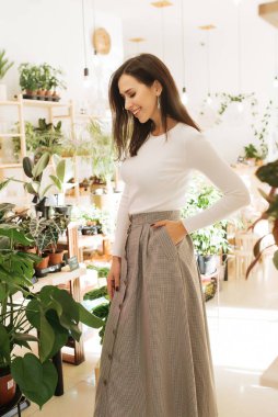 Genç zarif kız içeride poz veriyor. Duvarda ve pencerede zarif elbiseler içinde güzel bir iş kadını.