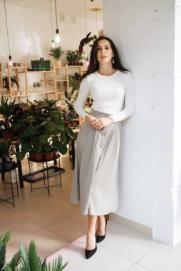 Genç zarif kız içeride poz veriyor. Duvarda ve pencerede zarif elbiseler içinde güzel bir iş kadını.