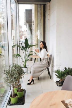 Genç zarif kız içeride poz veriyor. Duvarda ve pencerede zarif elbiseler içinde güzel bir iş kadını.