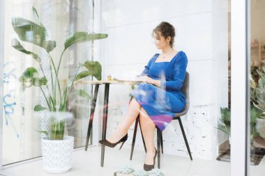 Genç zarif kız içeride poz veriyor. Duvarda ve pencerede zarif elbiseler içinde güzel bir iş kadını.