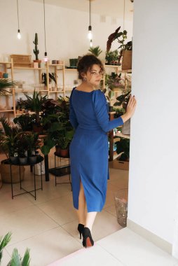 Genç zarif kız içeride poz veriyor. Duvarda ve pencerede zarif elbiseler içinde güzel bir iş kadını.