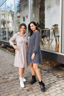 Genç zarif kız içeride poz veriyor. Duvarda ve pencerede zarif elbiseler içinde güzel bir iş kadını.