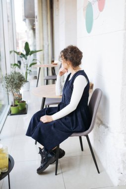 Genç zarif kız içeride poz veriyor. Duvarda ve pencerede zarif elbiseler içinde güzel bir iş kadını.