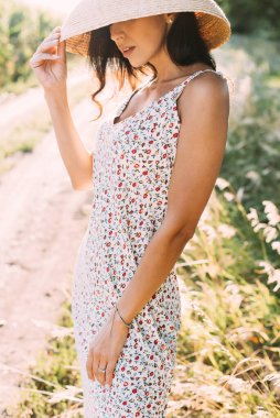 Zarif bir yaz elbisesi içinde genç ve güzel bir esmer ve güneş ışınlarında hasır şapkalı doğa yürüyüşünde.