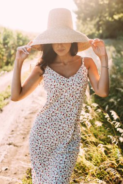 Zarif bir yaz elbisesi içinde genç ve güzel bir esmer ve güneş ışınlarında hasır şapkalı doğa yürüyüşünde.