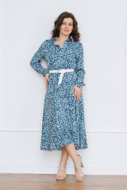 Yetişkin genç bir kadın, pencerenin yanındaki parlak bir odada güzel, mavi bir elbiseyle dinleniyor.