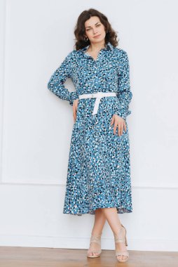 Yetişkin genç bir kadın, pencerenin yanındaki parlak bir odada güzel, mavi bir elbiseyle dinleniyor.