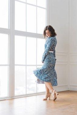 Yetişkin genç bir kadın, pencerenin yanındaki parlak bir odada güzel, mavi bir elbiseyle dinleniyor.