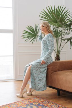 Genç zarif kız içeride poz veriyor. Duvarda ve pencerede zarif elbiseler içinde güzel bir iş kadını.