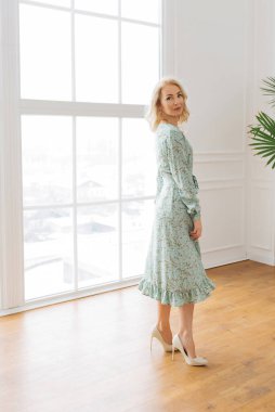 Genç zarif kız içeride poz veriyor. Duvarda ve pencerede zarif elbiseler içinde güzel bir iş kadını.