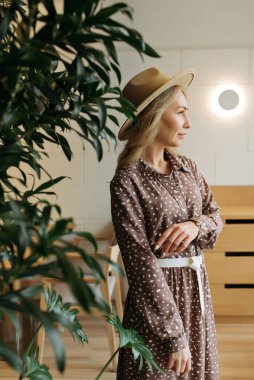 Yetişkin genç bir kadın, pencerenin yanındaki parlak bir odada güzel, mavi bir elbiseyle dinleniyor.