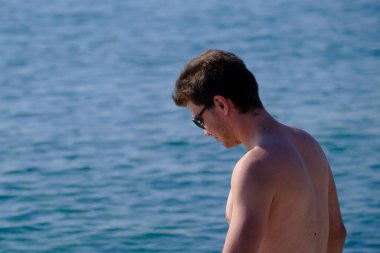 Deniz yüzeyinin arka planında 25-30 yaşlarında yakışıklı bir adamın portresi. Güneş gözlüklü, çıplak gövdeli, kumral, kalın saçlı.