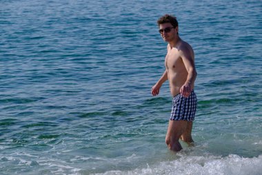 Deniz yüzeyinin arka planında 25-30 yaşlarında yakışıklı bir adamın portresi. Güneş gözlüklü, çıplak gövdeli, kumral, kalın saçlı.