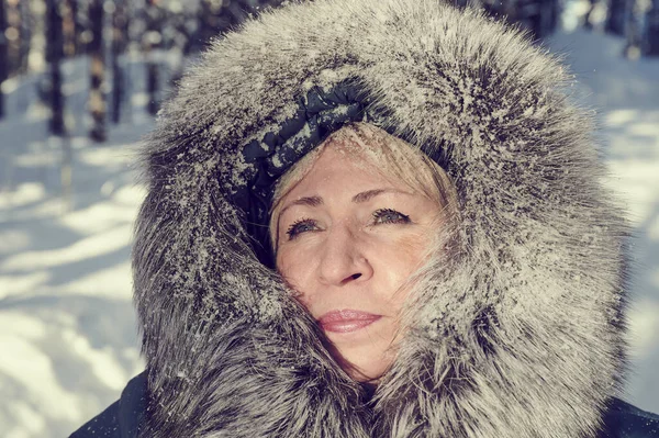 A 55-60-year-old blonde woman in winter clothes - a jacket with a polar fox edge under sunlight outdoors on the background of a snow-covered forest and snowfall.
