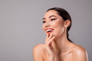Charming young brunette woman with flawless makeup and glowing skin posing against a gray background, showcasing elegance, self-confidence, and a sense of modern beauty and fashion.