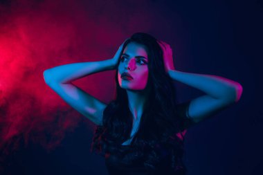 Stylish brunette model in a dark setting with pink and blue neon lighting, evoking modern beauty, elegance, and a glamorous nightlife atmosphere.