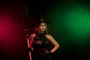 A bewitching woman dressed as a witch poses with a broom against a colorful foggy background, embracing Halloween magic and fantasy.