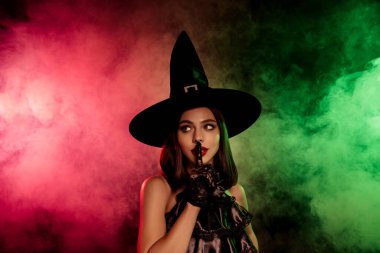 A mysterious and beautiful witch posing in a black hat surrounded by smoky red and green lights, conveying mystery and magic.