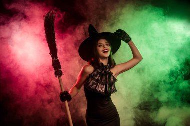 Charming portrait of a glamorous witch in a black outfit, holding a broomstick amidst colorful fog, evoking Halloween energy.