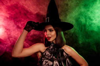 Stylish young woman wearing a classic witch costume with a pointy hat against a dramatic red and green misty background.