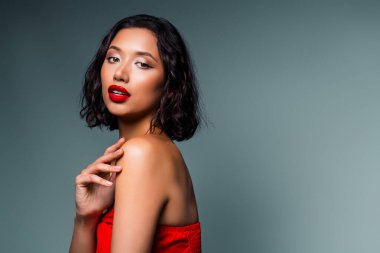 Kalın kırmızı dudaklı ve şık saçlı güzel esmer kadın zarif, cazibeli ve modern estetiği vurgulayan, minimalist gri arka plana karşı canlı bir şekilde poz verir.