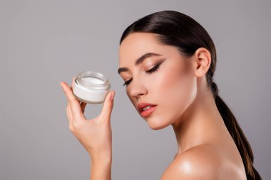 Yüz kremi kavanozu tutan esmer bir kadının yakın plan portresi. Cilt ve kozmetik ürünlerini temsil ediyor. Onun zarif ifadesi ve kusursuz teni lüks ve modaya uygun bir atmosferi geliştirir..