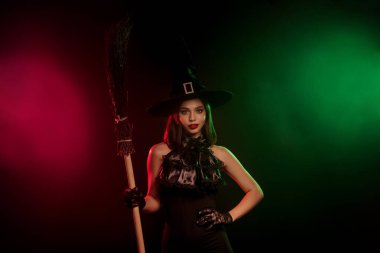 Cadılar Bayramı temalı, kırmızı ve yeşil sisli ışıklandırmalı canlı bir arka plana karşı elinde süpürge sopasıyla göz alıcı bir cadının esrarengiz tasviri..