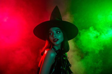 Cadı gibi giyinmiş güzel bir kadın. Şık bir şapka takıyor ve yeşil ve kırmızı sisin arasında duruyor, büyülü bir Cadılar Bayramı temasını çağrıştırıyor..