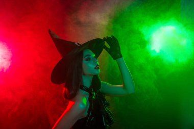 Cadı gibi giyinmiş şık genç bir kadın, kırmızı ve yeşil ışığın altında kendine güvenen bir sis içinde Cadılar Bayramı atmosferi yaratıyor..