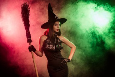 Büyüleyici Cadılar Bayramı temalı görüntüde şık giyinmiş büyüleyici bir cadı canlı dumanlı bir arka plana karşı güvenle poz veriyor, sihir ve gizemi çağrıştırıyor.