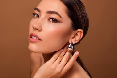 Zarif küpeler takan genç bir esmer kadının dikkat çekici yakın çekimi, kusursuz makyaj, pürüzsüz cilt, ve zarif bir stüdyoda sofistike bir bej arka plana karşı güzellik.