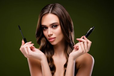 Göz kamaştırıcı esmer bir kadın elinde maskara aplikatörü ve yeşil bir arka planda duran kabıyla güzelliği, kozmetiği ve lüksü vurguluyor. Makyaj ve moda temaları için mükemmel.