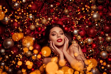 Çarpıcı bir model, kırmızı ve altın sarısı Noel süsleri ve parıldayan ışıklar arasında duruyor. Güzellik kampanyaları ve mevsimsel pazarlama malzemeleri için mükemmel bayram cazibesi, zerafet ve şenlik havası yayıyor..