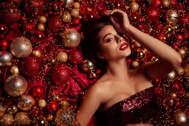 Parlak kırmızı elbiseli güzel bir model altın ve kırmızı takılar ve peri ışıkları arasında uzanır, neşeli tatil cazibesini, lüks moda cazibesini ve Noel ve yeni yıl kampanyaları için şenlikli atmosferi çağrıştırır..